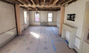 A partially renovated room with exposed wooden ceiling beams, large windows allowing natural light, a fireplace on one wall, and a covered floor protected by paper and blue tape. The walls are prepped for painting or further construction work.