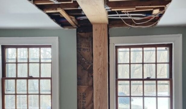  A partially renovated interior with two large windows featuring wooden frames, separated by an exposed wooden beam. The ceiling above the beam is open, revealing insulation, wiring, and structural elements, indicating ongoing construction or repair work.